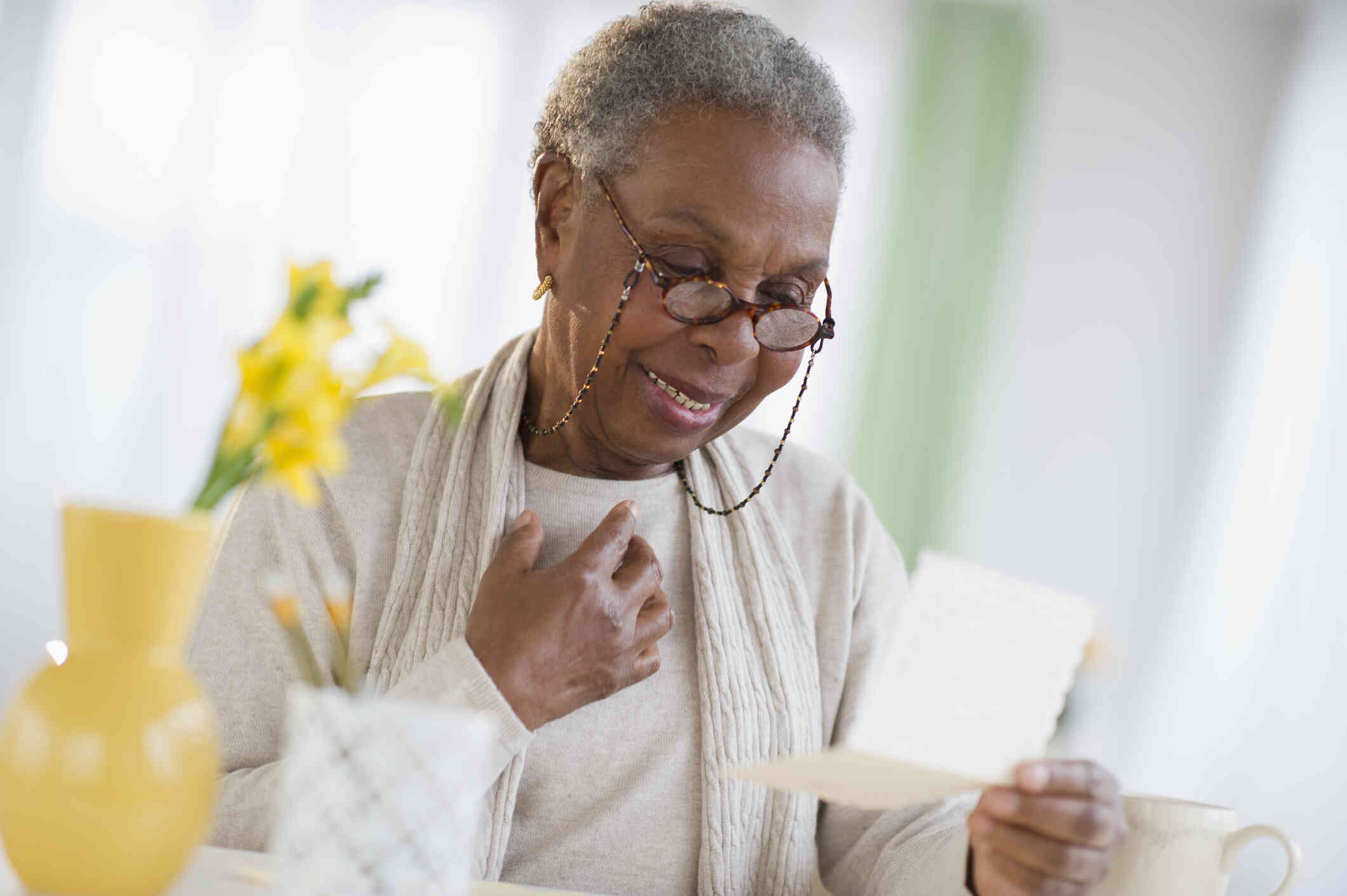 An elderly woman with glasses stands in her home and smiles down at a peice of paper in her hand.